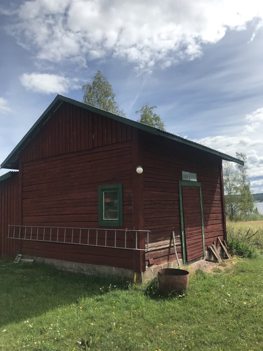 Ett rött trähus som tidigare varit stall med grönt fönster, dörr och gräsmark i förgrunden.