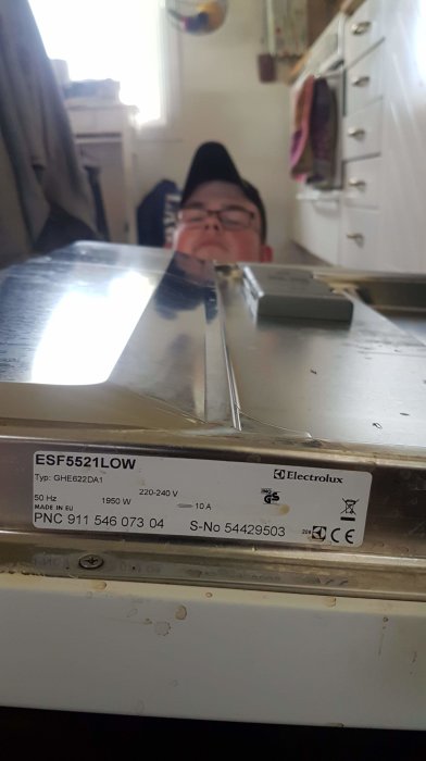 En person speglar sig i en diskmaskins lucka med en specifikationsetikett från Electrolux synlig i förgrunden.