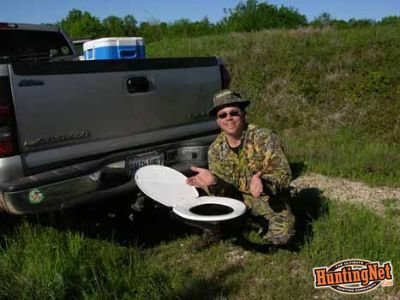 Person squatting next to a portable 'bumper dumper' toilet attached to the back of a pickup truck in an open field.