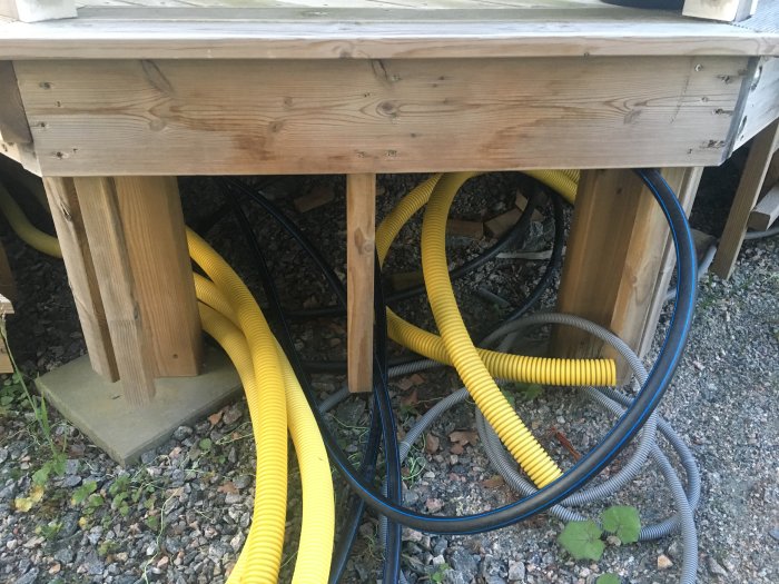 Oorganiserade gula och gråa PEM-slangar och flexrör under en träkonstruerad altan, intill en betongplatta.