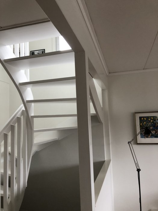 Vitmålad trappa med öppet koncept, vitmålade väggar och en golvlampa till höger.