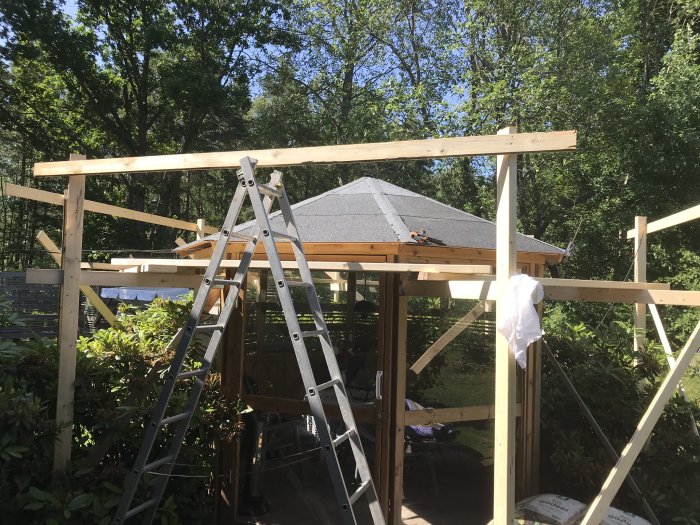 Trästomme av byggprojekt med presenning på tak, stegar och byggmaterial omkring.