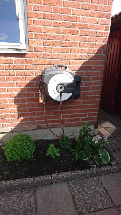 En nyinstallerad automatisk slangvinda på en tegelhusfasad vid en blomsterrabatt.