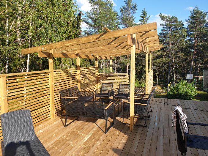 Trädäck med pergola och sittgrupp, omgivet av gröna träd och skog i bakgrunden.