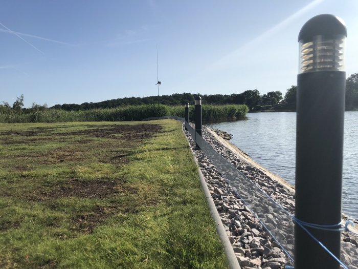 Gräsmatta vid vattenkanten med ojämn grästillväxt och nätförseglad stenrad längs en sjö.