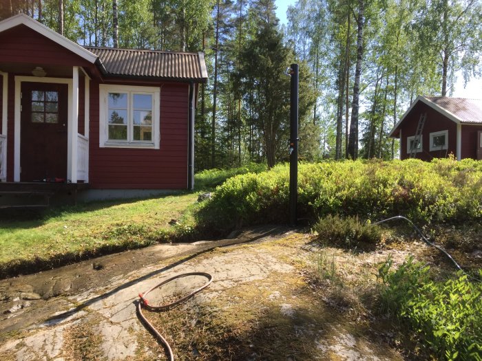 Berghäll i trädgården framför röda stugor, omgiven av grönska under en solig himmel.