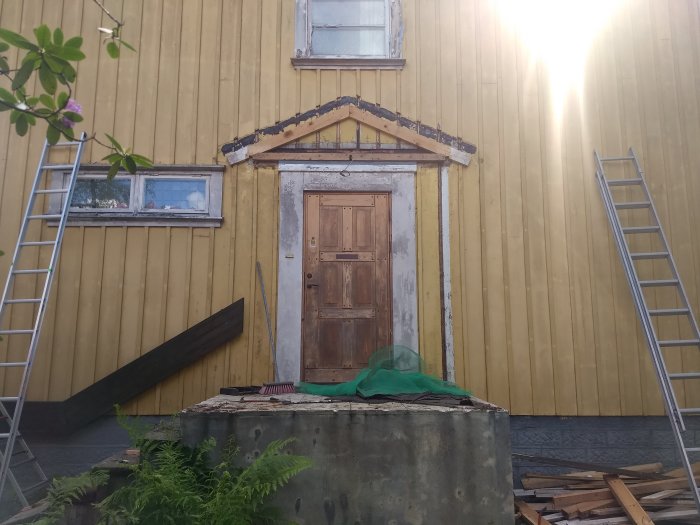 Rivningsarbete runt en entrédörr till ett gult hus, förbereder för nytt entrétak med materialrester och stegar synliga.