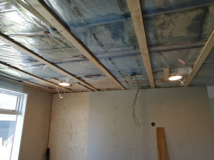 Interiör av ett rum under renovering med isolering i taket och två provisoriska lampor tända.