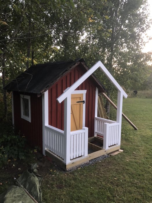 Röd lekstuga med orenoverat svart tak och nybyggd vit farstukvist, omgiven av grönska.