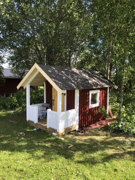 Röd lekstuga med nytt mörkgrått tak och vit farstukvist omgiven av gröna träd och gräsmatta.