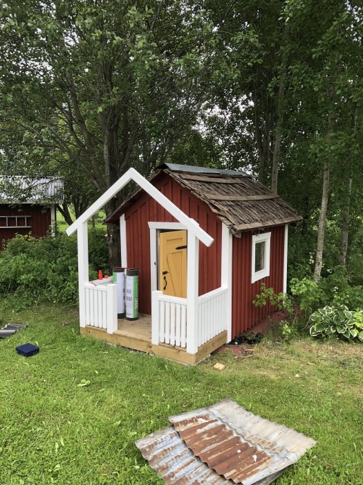 Rödmålad lekstuga med ofärdigt tak och ny farstukvist, rostiga plåtar ligger på marken.