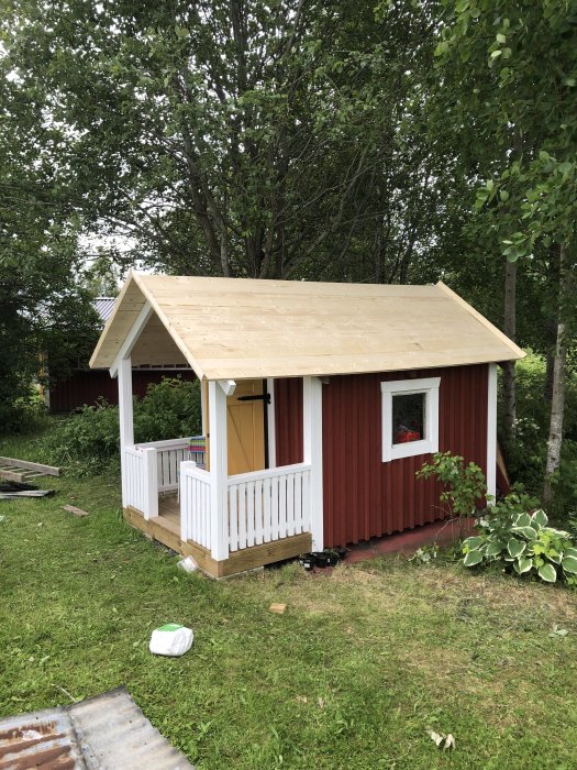 Nybyggt oskyddat tak på en röd lekstuga med farstukvist, redo för plåtläggning, omgiven av grönska.