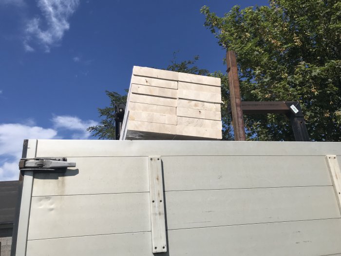 Nyligen uppsatt balk till garageport och staplade hammarband som väntar på att resas på ett takbygge, med himmel och träd i bakgrunden.
