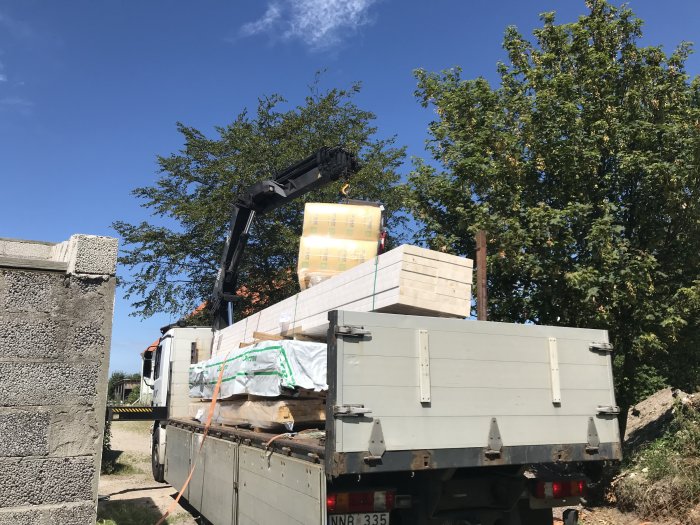 Lastbil med kran lyfter byggmaterial såsom takreglar för hemmaprojekt mot en bakgrund av gröna träd.