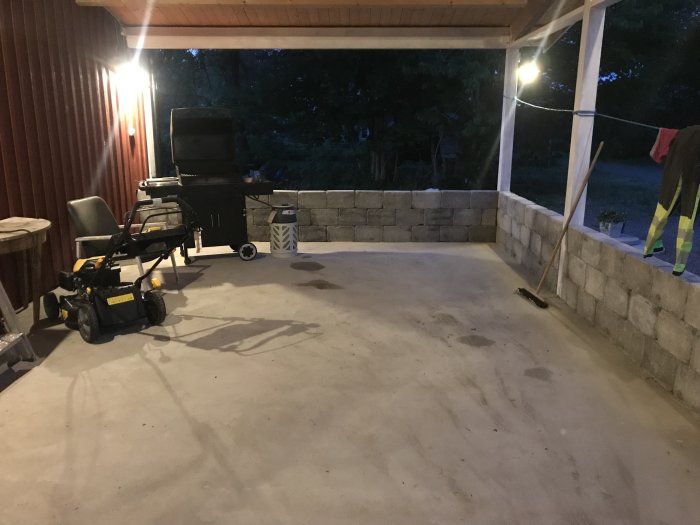 Nygjuten betongplatta under tak med grill, trädgårdsutrustning och belysta träväggar i skymningen.
