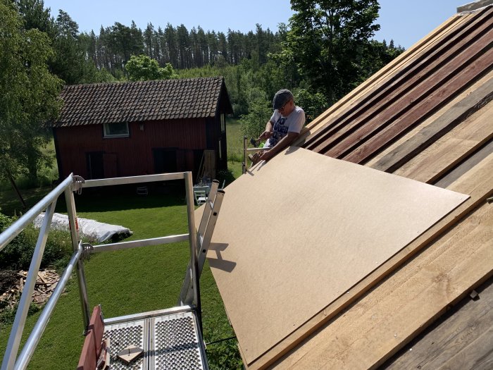 En person som monterar board på ett takstomme med en röd stuga och grönska i bakgrunden.