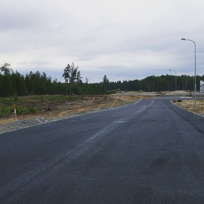 Tomtmarkerad för bygge med grus och pålar längs en asfalterad väg och skog i bakgrunden.