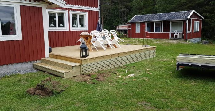 Nybyggd träaltan med bord och vita stolar framför rött hus, i väntan på fler sittplatser.