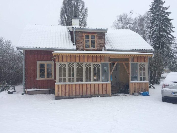 En äldre rödmålad trävilla med snötäckt tak och en pågående tillbyggnad av en glasveranda, omgiven av ett snöigt landskap.