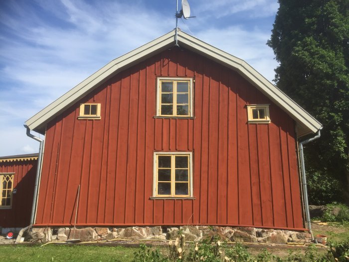 Rödmålad traditionell svensk lada med vita fönsterkarmar och stengrund, omgiven av grönska.