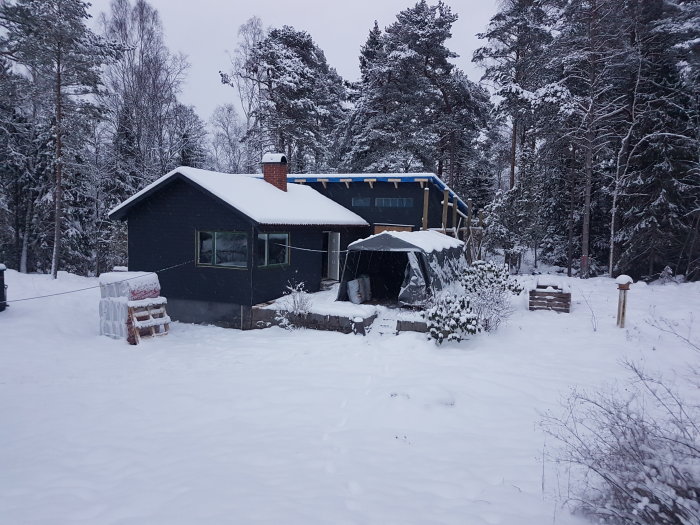 Ett hus och en förrådsbyggnad täckta med snö bland träd i ett snölandskap.