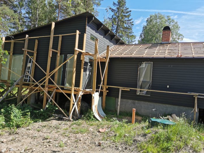 Byggställningar bredvid ett svart trähus under renovering med oavslutad takläggning och byggmaterial på marken.