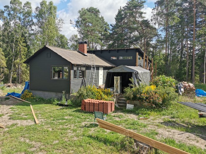 Ett pågående byggprojekt av ett hus med skogsbakgrund och byggmaterial spridda på gräsmattan.