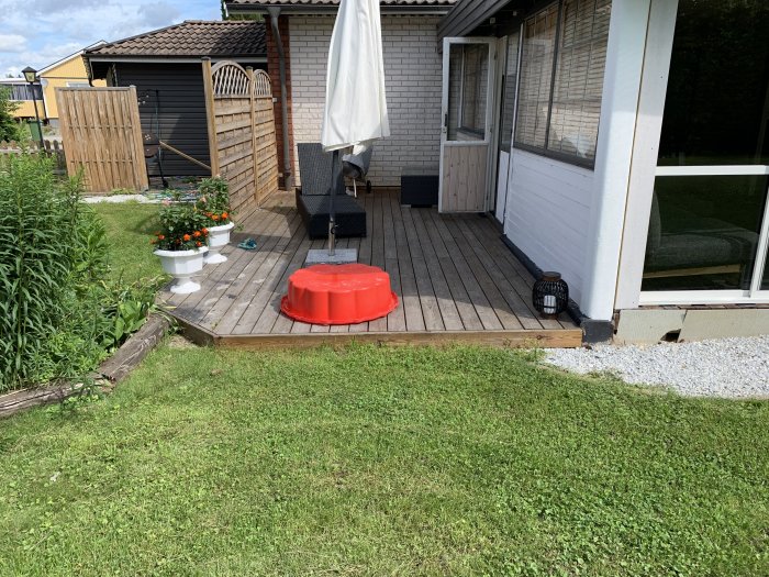Existerande trädäck vid ett hus med gräsmatta, möbler och en röd leksak, redo för utbyggnad.