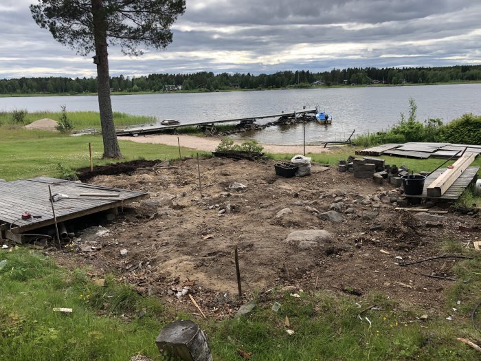 Riven byggplats för strandstuga med sand/skogsmark och byggmaterial vid vattnet.