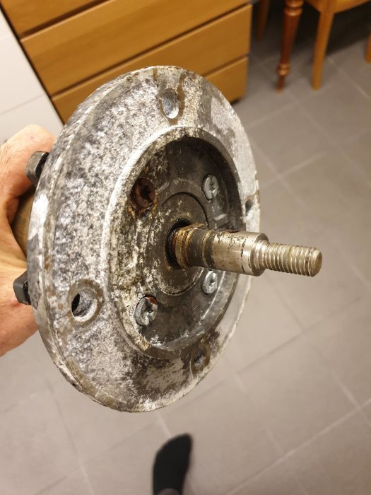 En hand håller en sliten elektrisk motoraxel med synlig gänga och korrosion mot en inomhusbakgrund.