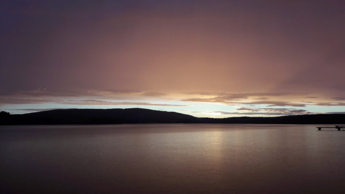 Solnedgång över ett stilla vatten med lila himmel och mörka silhuetter av kullar.