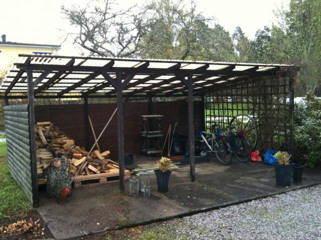 Åpen vedskjul med trestolper, sykkel, vedstabler, og diverse redskaper. Skjulet har tak av plastplater og åpen front.