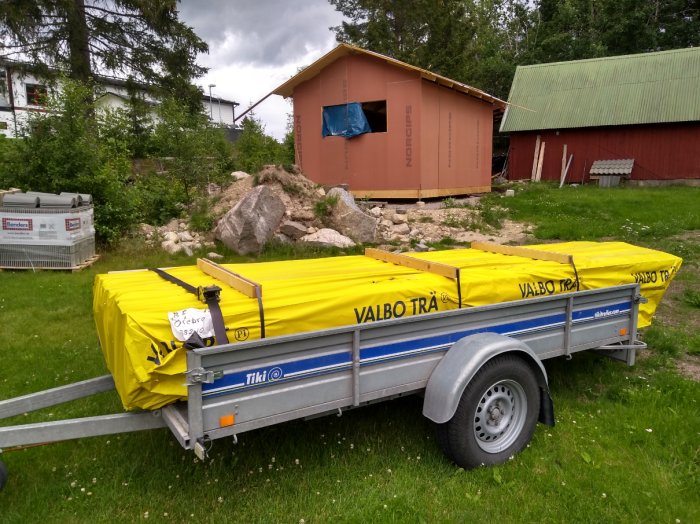 Släpvagn lastad med gula presenningsskyddade paket av ytterpanel framför oavslutat husbygge.