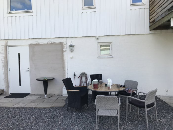 Husfasad med ett igensatt fönsterområde, vit ytterdörr och uteplats med möbler.