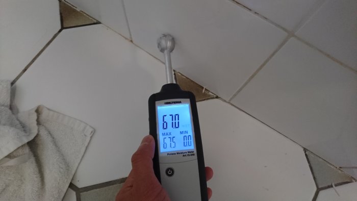 Hand håller en fuktmätare mot en kaklad vägg, indikerar hög fuktighet med siffran 60 och texten "WET".