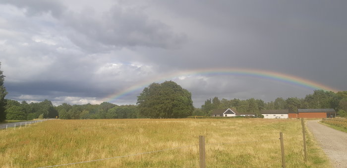 En regnbåge över ett landskap med öppet fält, träd och gårdshus under en dramatisk himmel.