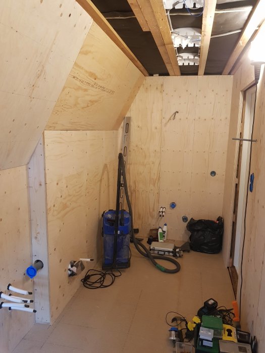 Badrumsrenovering med nyinstallerad plywood på väggar och snedtak, elinstallation utspridd och byggverktyg på golvet.