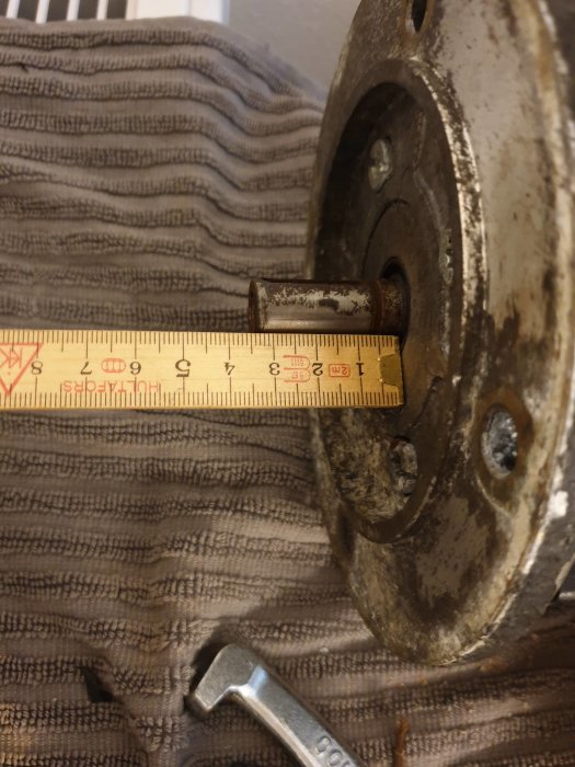Mätning av en 14 mm motoraxel med skjutmått på en gammal motor, rost och verktyg syns.