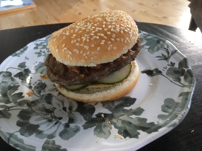Hamburgare med sesambröd och inlagd gurka på blommigt fat.