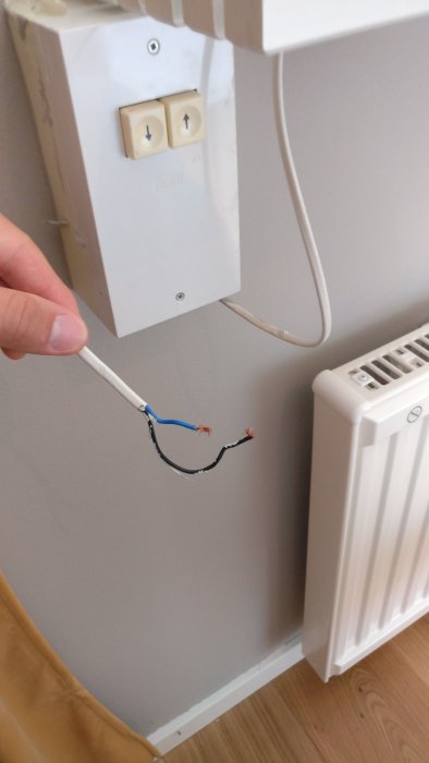 En hand håller en avskalad elektrisk kabel med exponerade ledare framför en brytare och längs en vägg.