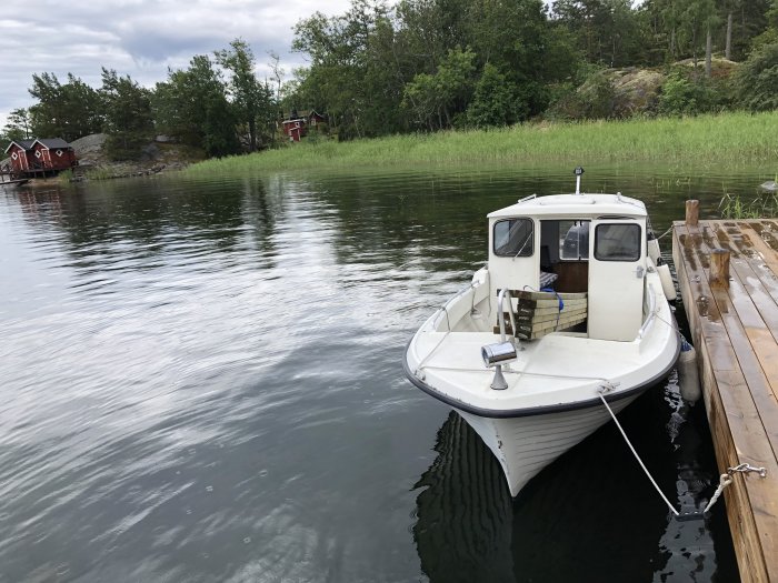 Vit motorbåt förtöjd vid en träbrygga med röda stugor och skog i bakgrunden.