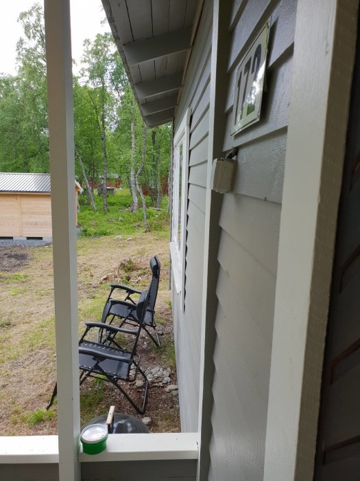 Utsikt från husveranda med stolar, skog i bakgrunden och husnummer vid dörren.