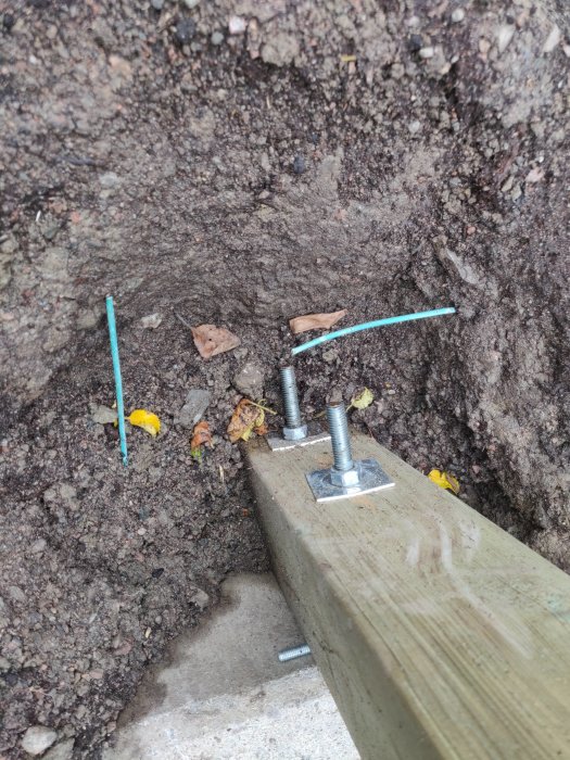 M12 gängstång och L-stöd i betong med 16cm mellanrum stabiliserar en trästolpe i marken.