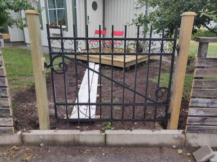 En svart smidesgrind mellan två trästolpar vid en trädgårdsgång, närbild på monteringsdetaljer.