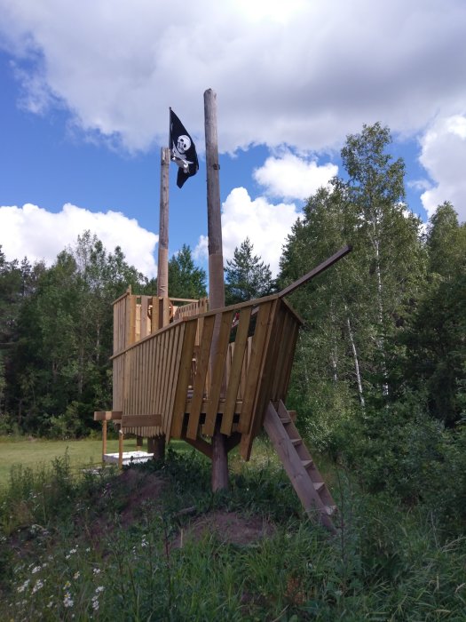 Trädkoja med piratflagga på hög mast under blå himmel omgiven av grönska.