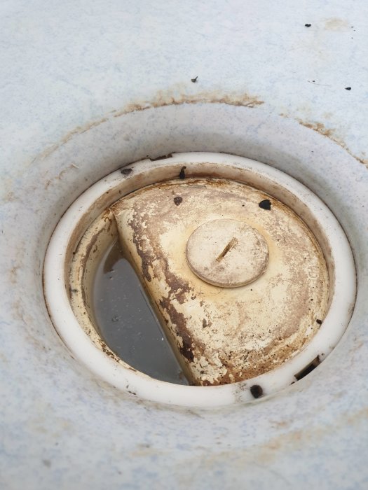En smutsig och rostig golvbrunn med en lossnad övre ring och synliga rester av rostade skruvar.