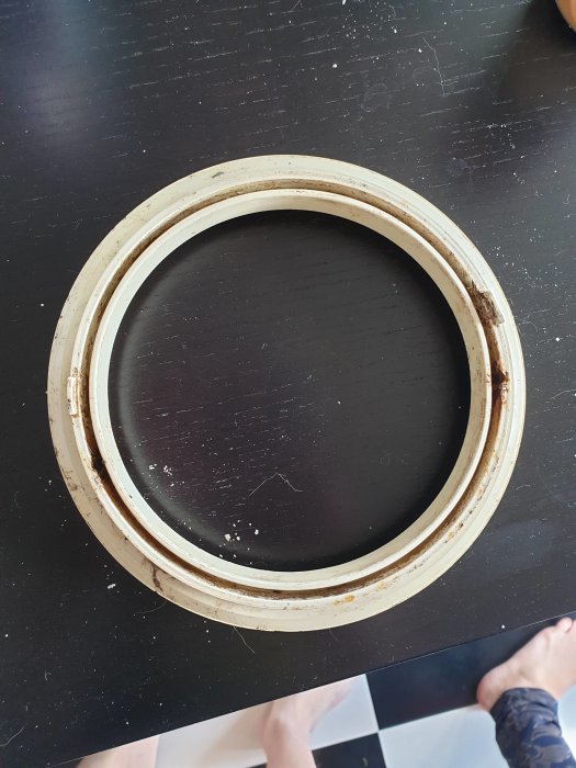 Rostig övre ring till golvbrunn på ett svart underlag med personens fötter i bakgrunden.