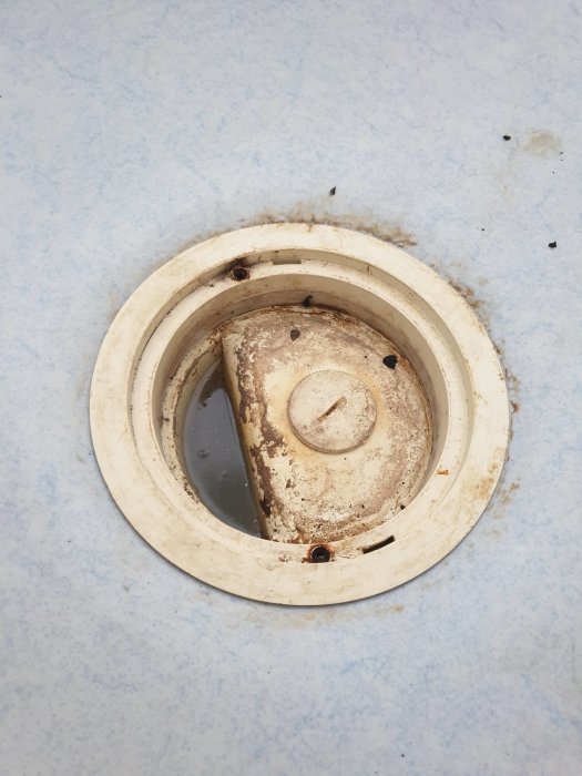 Rostad och smutsig golvbrunn med en lossad övre ring och rostiga skruvar på ett kaklat badrumsgolv.