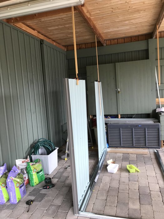 Pågående montering av ett grått plåtförråd inne i en carport med verktyg och byggmaterial utspridda på marken.