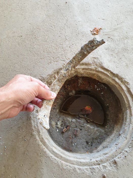 Hand som lyfter ett slitet galler från ett garageavlopp med synlig distansring och smutsigt vatten.